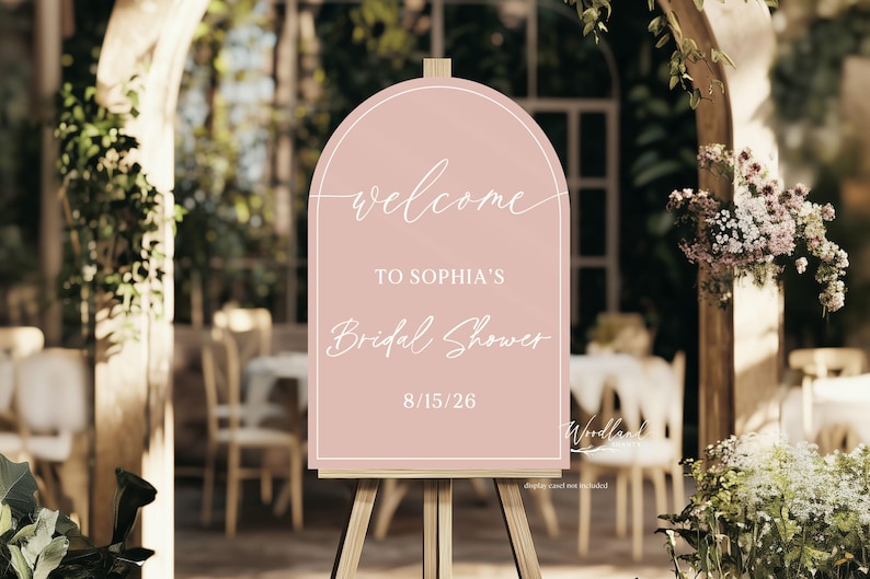 May include: A pink sign on a wooden easel, with the words "Welcome to Sophia's Bridal Shower" and the date 15/08/26. The sign has an arched top and is set against a blurred outdoor background.