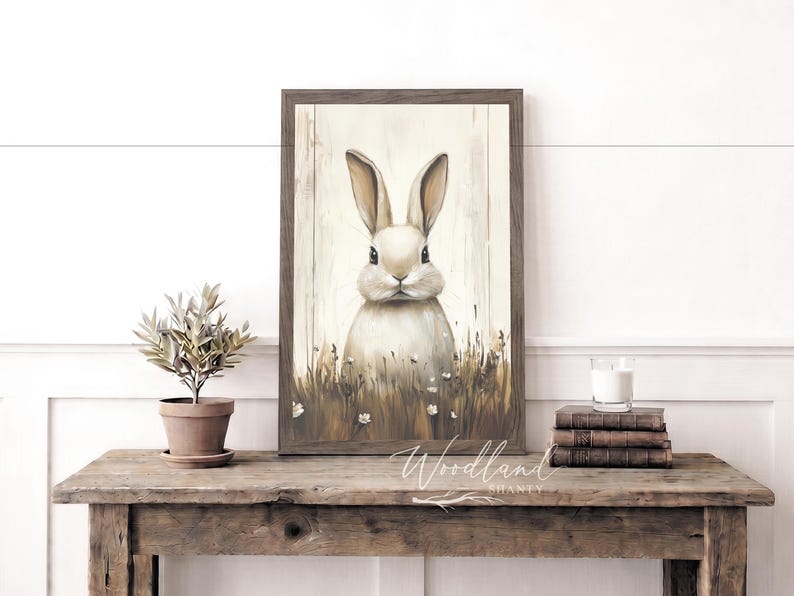 May include: A framed print of a rabbit in a field of grass and flowers. The artwork has a rustic, vintage style with a neutral color palette. The frame is a dark brown color. The print is displayed on a wooden table.