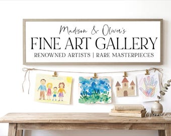 May include: A rectangular wooden-framed sign with white background and black text reading Madison & Oliver's FINE ART GALLERY RENOWNED ARTISTS | RARE MASTERPIECES. Below, a string with clips holds four children's drawings of a family, landscape, castle, and handprint.