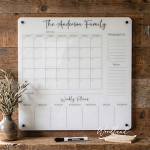 May include: A white acrylic family calendar with the text "The Anderson Family" at the top. It features a monthly calendar, a shopping list, and weekly plans. A dry-erase marker and a small stack of paper are on the wooden surface.