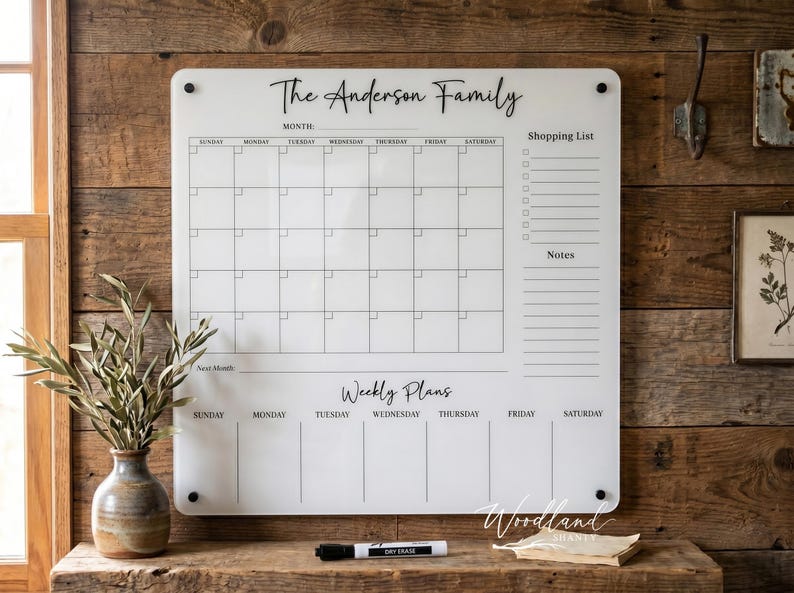 May include: A white acrylic family calendar with the text "The Anderson Family" at the top. It features a monthly calendar, a shopping list, and weekly plans. A dry-erase marker and a small stack of paper are on the wooden surface.