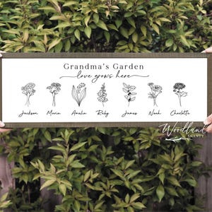 May include: A framed wooden sign with the text "Grandma's Garden love grows here." The sign features line drawings of flowers and names: Jackson, Maria, Amelia, Ruby, James, Noah, and Charlotte. The sign is held by two people.