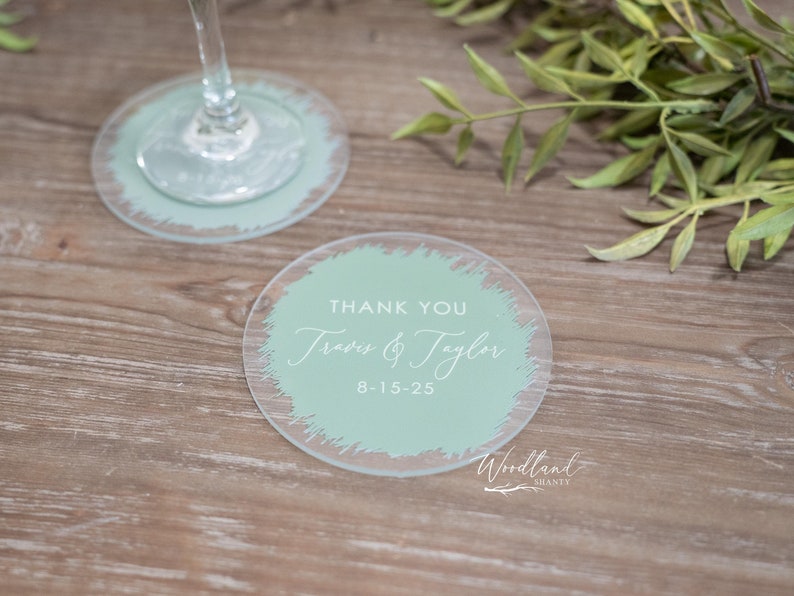 May include: Two round, clear glass coasters with a light green, brushstroke design. The top coaster has the text "Thank You Travis & Taylor 8-15-25" and the text "Woodland Shanty" at the bottom.