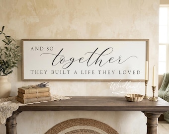 May include: A rectangular wooden sign with the words "AND SO together THEY BUILT A LIFE THEY LOVED" in black script. The sign is displayed above a dark wooden table with decorative items, including books, candles, and a small bowl.
