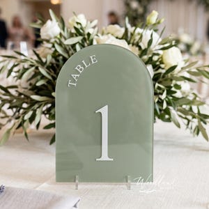 May include: Sage green table number with the word "TABLE" arched at the top and the number "1" in the center. The table number is displayed on a white tablecloth with a floral arrangement in the background.