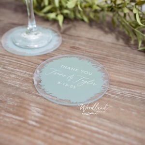 May include: A round, light blue glass coaster with a white brushstroke design. The text "Thank You" is written in white script, followed by "Jess & Tyler" and the date "8-15-25". The text "Woodland" is written in white script below the design.