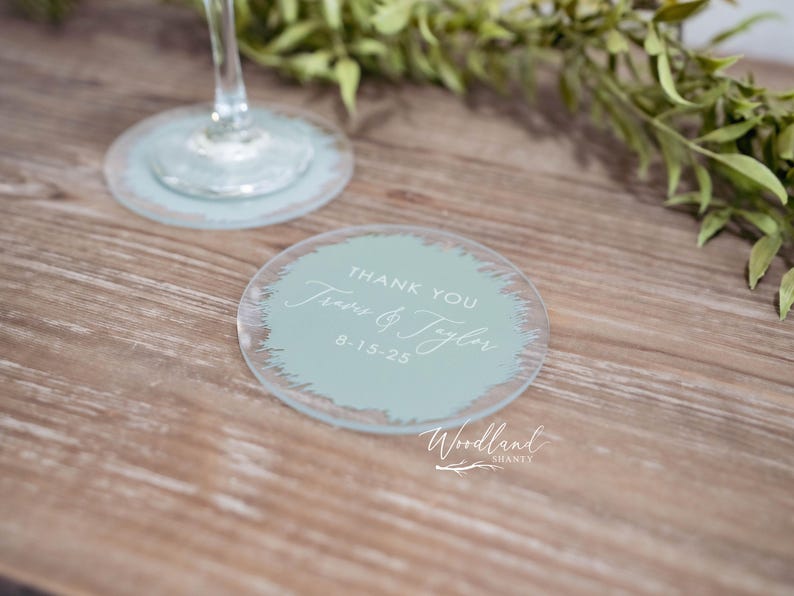 May include: A round, light blue glass coaster with a white brushstroke design. The text "Thank You" is written in white script, followed by "Jess & Tyler" and the date "8-15-25". The text "Woodland" is written in white script below the design.