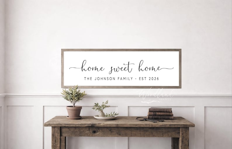 May include: A rectangular wooden sign with a white background and a brown frame. The sign reads "home sweet home" in elegant cursive, with "THE JOHNSON FAMILY - EST 2026" below. The sign is above a wooden table with plants and books.