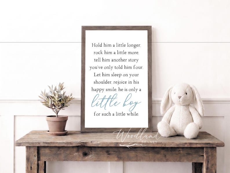 May include: A framed print with the text "Hold him a little longer...little boy for such a little while." A stuffed bunny and a potted plant sit on a rustic wooden table. The frame is dark brown.
