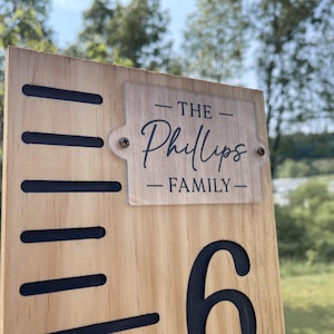 May include: Wooden growth chart with black markings and the number 6. A clear rectangular plaque is attached with the text "THE Phillips FAMILY". The chart is made of light-colored wood and is designed to measure height.