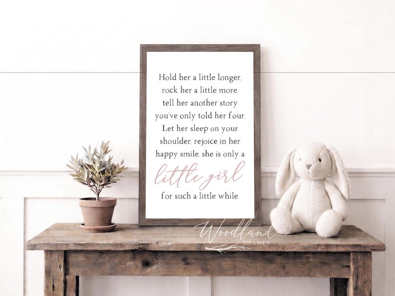 May include: A framed print with the text "Hold her a little longer" and a plush bunny on a wooden table. A small potted plant is also on the table. The print has a brown frame and the text is in black and pink.