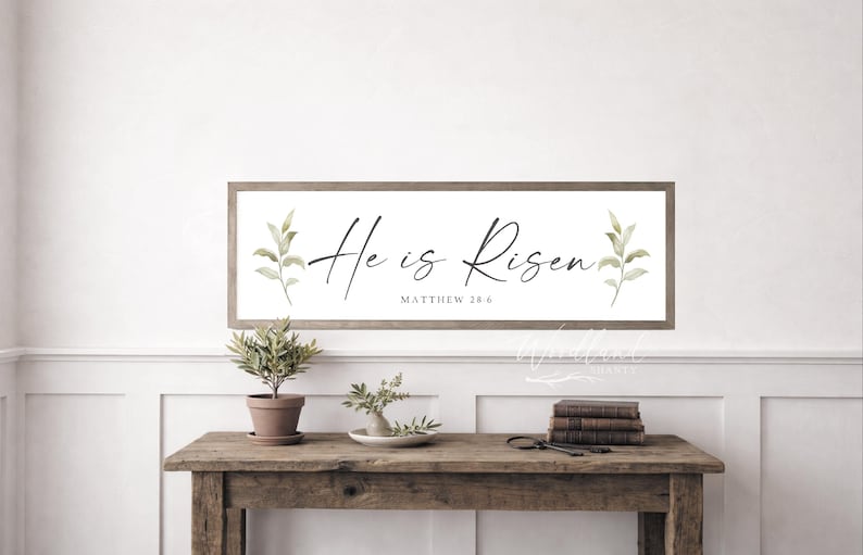Op de afbeelding: Een rechthoekig houten bord met de woorden "He is Risen" in elegant schrift, met de tekst "Matthew 28:6" eronder. Het bord is versierd met aquarel bladeren. Het bord wordt boven een houten tafel weergegeven.