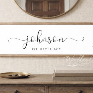 May include: A framed wooden sign with the name "johnson" in elegant cursive script. Below the name, the text reads "EST MAY 15, 2027". The sign is displayed above a wooden dresser, with a decorative tray and books.
