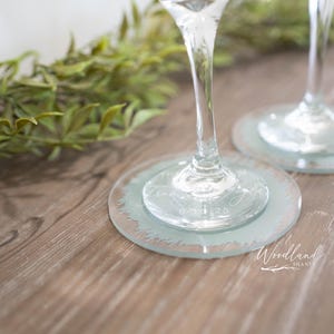 May include: Two clear glass wine glasses with etched designs on the bottom of the glass. The glasses are on light blue coasters with a decorative edge. The coasters are on a wooden surface.