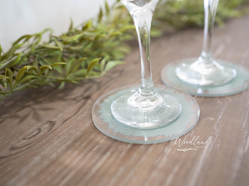 May include: Two clear glass wine glasses with etched designs on the bottom of the glass. The glasses are on light blue coasters with a decorative edge. The coasters are on a wooden surface.