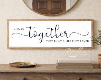 May include: A framed sign with the words "And so together they built a life they loved" in black script. The sign is displayed above a wooden console table with decorative items, including a small plant and a gold-toned tray.