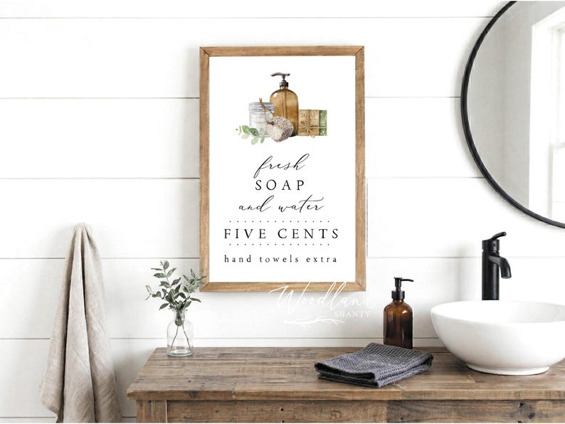 May include: A framed sign with the words "fresh soap and water" and "five cents hand towels extra." The sign is above a wooden vanity with a soap dispenser, a folded gray towel, and a white sink. A round mirror hangs on the wall.