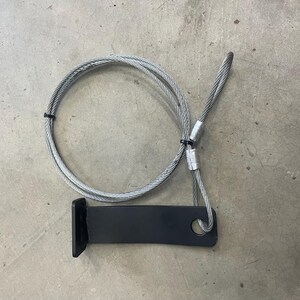 May include: A black metal bracket with a hole and a silver cable loop. The cable is secured with a black plastic tie. The bracket has a right angle bend.
