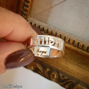 May include: A silver band ring featuring cut-out Hebrew lettering along the outer surface. The interior of the ring is engraved with the English word hope in a lowercase script font. The metal has a polished, reflective finish throughout.