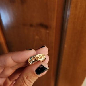 May include: A gold-colored band ring featuring a series of cut-out rectangular shapes along the outer surface. The ring has a polished metallic finish and a consistent width throughout its circular design, showcasing a repeating geometric pattern.
