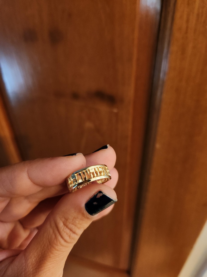 May include: A gold-colored band ring featuring a series of cut-out rectangular shapes along the outer surface. The ring has a polished metallic finish and a consistent width throughout its circular design, showcasing a repeating geometric pattern.