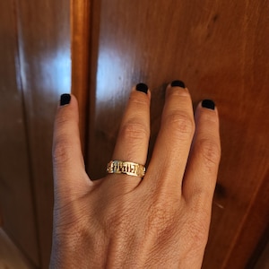 May include: A gold-toned band ring featuring a series of cut-out Hebrew characters encircling the entire circumference of the metal band. The ring has a polished finish and a uniform width, displaying distinct letter forms integrated into the design.