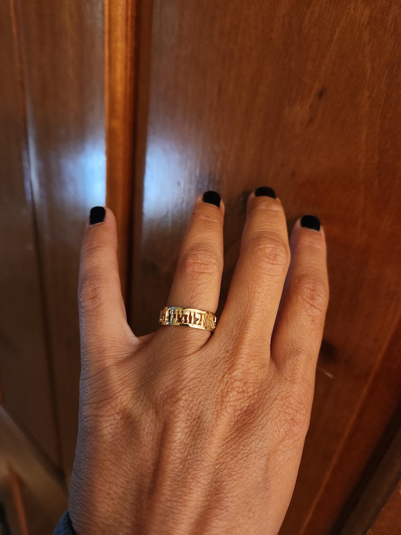 May include: A gold-toned band ring featuring a series of cut-out Hebrew characters encircling the entire circumference of the metal band. The ring has a polished finish and a uniform width, displaying distinct letter forms integrated into the design.