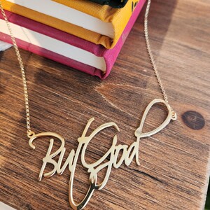 May include: A gold-toned metal necklace featuring a script-style pendant that reads "But God". The pendant is attached to a delicate, thin gold-toned chain. The lettering is connected in a flowing, cursive font with a polished, reflective finish.