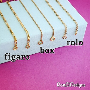 May include: Close-up of five different gold chain styles: figaro, box, rolo, cable, and curb. The chains are laid out on a light blue surface with the names of each chain style labeled below.