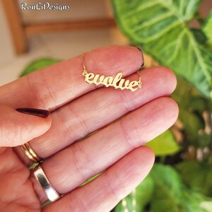 May include: A gold-colored necklace with the word "evolve" in cursive script. The necklace is held in a hand with a dark red nail polish. The background includes green foliage and a wooden surface.