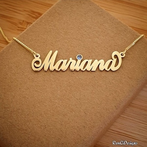 May include: A gold-colored necklace with the name "Mariana" in a cursive font. A small, round, blue gemstone is set above the letter "a". The necklace rests on a brown surface.