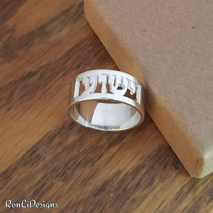 Könnte beinhalten: Ein silberner Ring mit dem hebräischen Wort "שלום" (Shalom), das aus der Band ausgeschnitten ist.