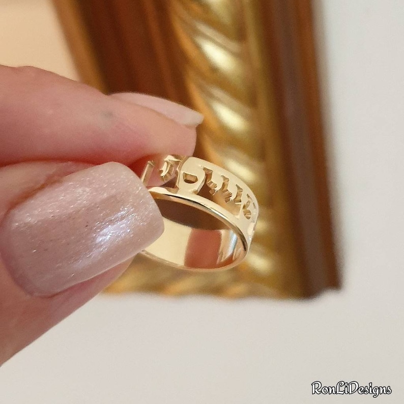 May include: A polished gold ring featuring a band with cut-out Hebrew lettering along the top edge. The metal has a reflective, smooth finish, and the design consists of a continuous openwork pattern where the characters form the structure of the ring.