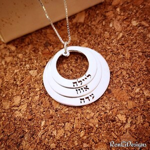 May include: Silver necklace with a layered circle pendant featuring Hebrew text. The pendant has three circles, each with a different Hebrew word. The words are "Chava", "Avigail", and "Hadar".