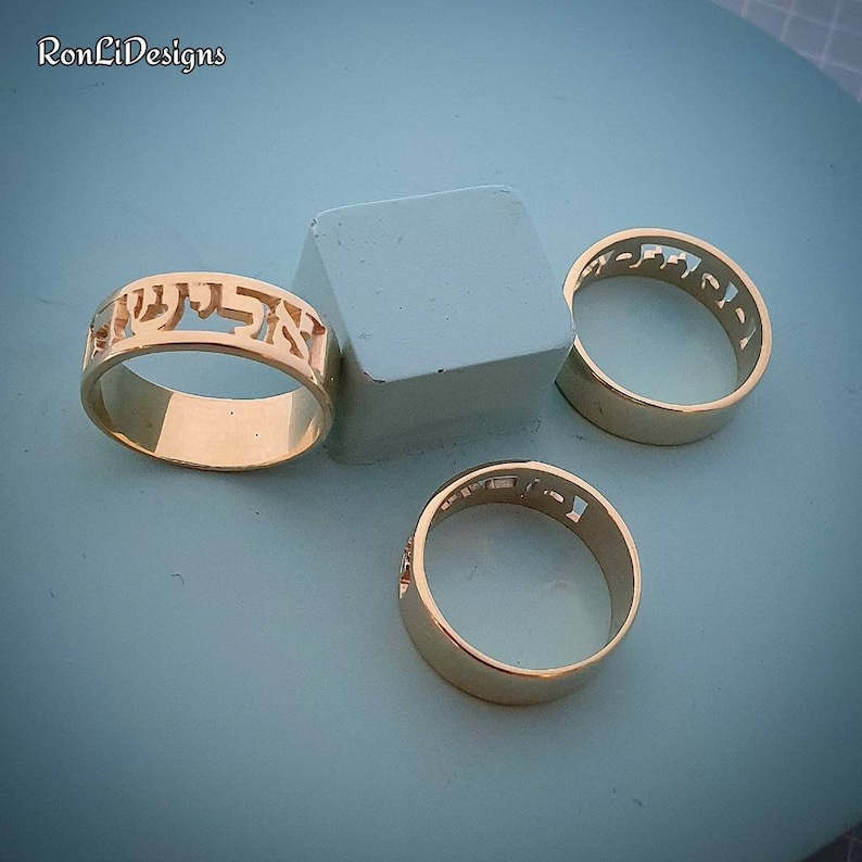 May include: Three gold-toned rings featuring cut-out Hebrew lettering along the band. The rings are arranged around a small, light blue square block. The text "RonLiDesigns" is visible in the upper left corner of the image.