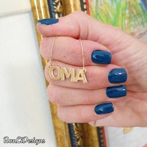 May include: Gold necklace with the word "OMA" spelled out in block letters. The necklace is being held by a hand with blue nail polish.