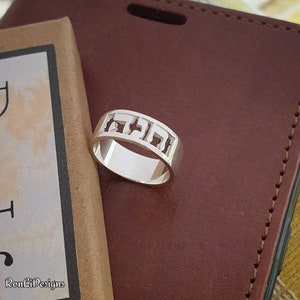 May include: A silver ring with a Hebrew inscription cut out of the band. The ring is on a brown leather surface.