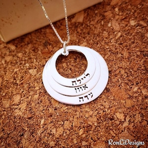 May include: Silver necklace with a layered circle pendant featuring Hebrew text. The pendant has three circles, each with a different Hebrew word. The words are "Chava", "Avigail", and "Hadar".