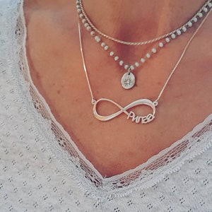 May include: A silver infinity necklace with the word "PWIB" inscribed on it. The necklace is layered with a silver chain and a beaded chain with a small silver sun pendant.