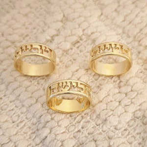 May include: Three gold-toned metal rings, each featuring a band with an openwork design consisting of Hebrew characters. The rings have a polished, reflective surface and a consistent circular shape, showcasing a cutout style along the upper portion.