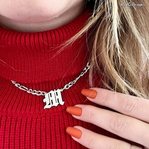 Op de afbeelding: Een zilverkleurige ketting met een enkele letter M-hanger in een gotisch lettertype. De hanger is bevestigd aan een metalen ketting met langwerpige ovale schakels, rustend tegen een felrode geribbelde stof aan.