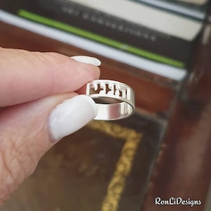 May include: A polished silver band ring featuring a cutout design of Hebrew characters along the top section. The metal has a smooth, reflective finish, and the openwork lettering creates a distinct pattern that wraps around the upper portion of the ring.