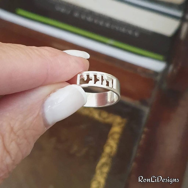 May include: A polished silver band ring featuring a cutout design of Hebrew characters along the top section. The metal has a smooth, reflective finish, and the openwork lettering creates a distinct pattern that wraps around the upper portion of the ring.
