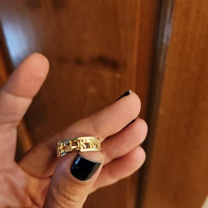 May include: A gold-colored metal ring featuring cut-out Hebrew lettering around the entire band. The ring has a polished, reflective surface and a continuous design where the characters form the structure of the band itself.