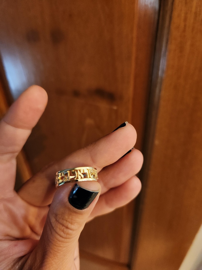 May include: A gold-colored metal ring featuring cut-out Hebrew lettering around the entire band. The ring has a polished, reflective surface and a continuous design where the characters form the structure of the band itself.