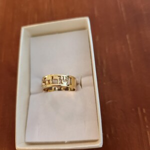 May include: A gold band ring featuring cut-out Hebrew lettering along the circumference. The ring is presented inside a rectangular white box with a soft interior lining, showcasing the polished metallic finish and the openwork design of the characters.