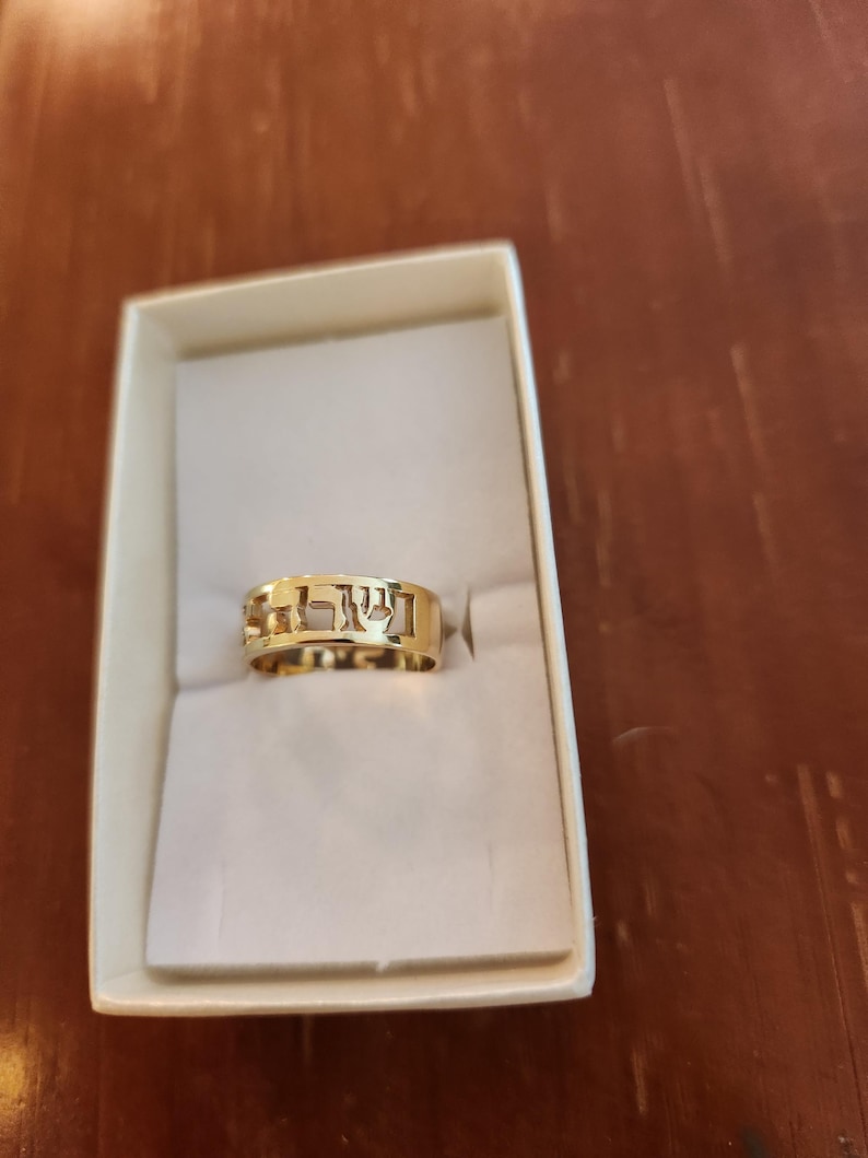 May include: A gold band ring featuring cut-out Hebrew lettering along the circumference. The ring is presented inside a rectangular white box with a soft interior lining, showcasing the polished metallic finish and the openwork design of the characters.