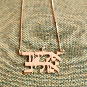 May include: A rose gold-toned necklace featuring a pendant with Hebrew text. The pendant is suspended from a delicate box chain. The Hebrew characters are arranged in two lines, creating a solid, block-like design with a polished, reflective surface.