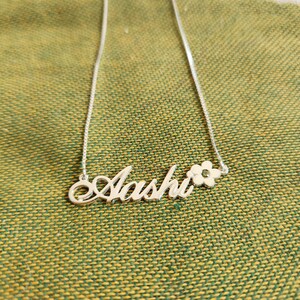 May include: A silver-toned necklace featuring a cursive nameplate that reads Aashi, with a small five-petaled flower charm attached to the right side of the name. The pendant is suspended from a thin, delicate silver-toned box chain.