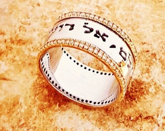 May include: A silver ring with gold accents and embedded gemstones. The ring features Hebrew text around the band. The ring is set against a textured, light brown surface.
